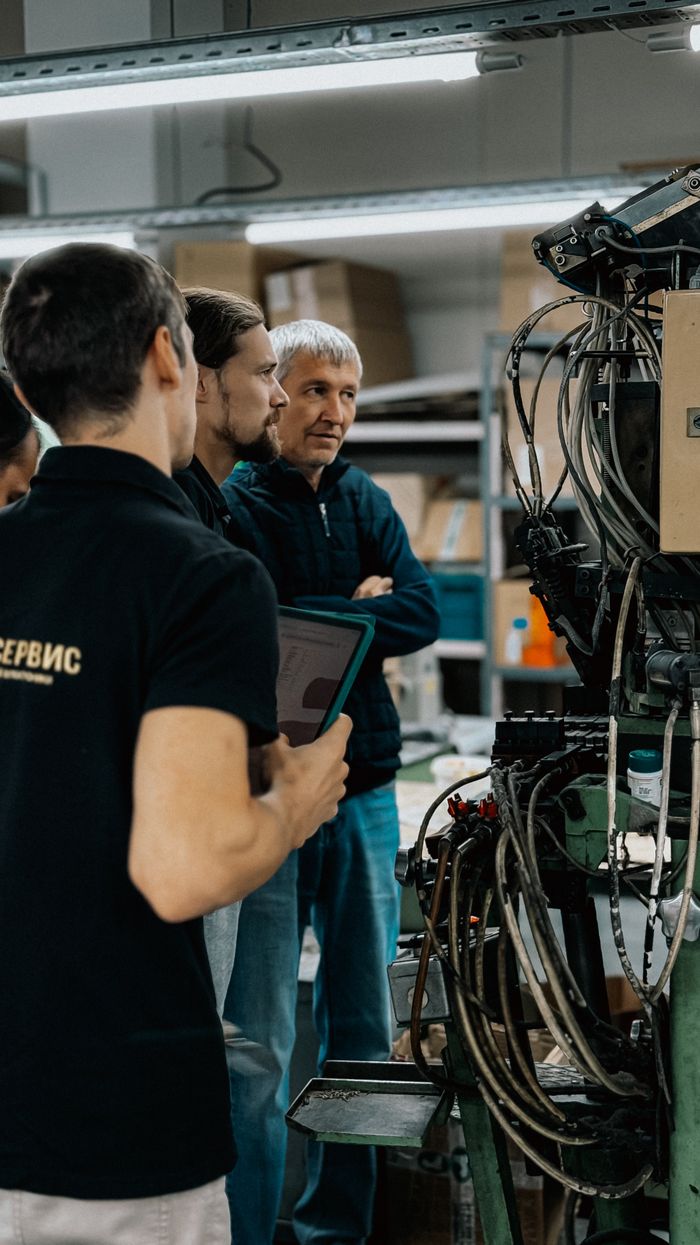 About Three adults engaged in a discussion around complex machinery in a factory environment.